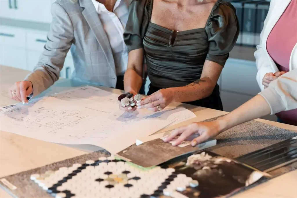Three people review floor plans and design samples at a table in a well-lit modern kitchen, discussing materials like tiles and fabric swatches, highlighting the importance of feeling a space vs. seeing it.