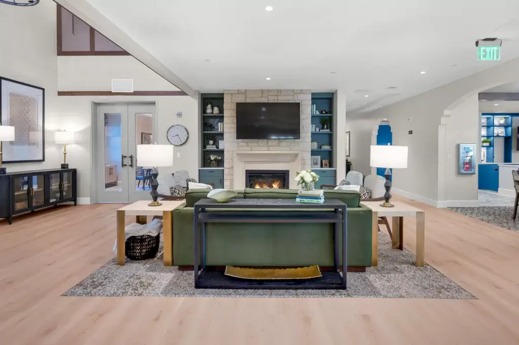 Modern living room with a green sofa, lamps on side tables, a fireplace with a TV above it, built-in shelves, light wood flooring, and neutral decor in soft, calming tones.