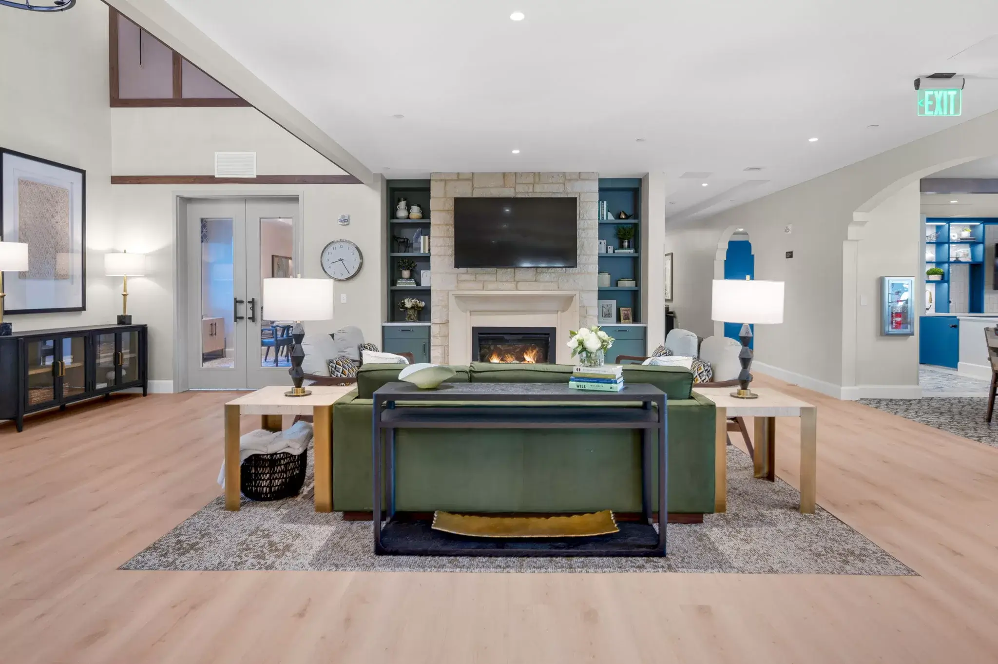 Modern living room with a green sofa, lamps on side tables, a fireplace with a TV above it, built-in shelves, light wood flooring, and neutral decor in soft, calming tones.