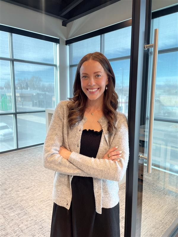 A woman with long brunette hair, wearing a grey sweater and black dress, stands smiling against a modern, stylish glass door with a conference room in the background.