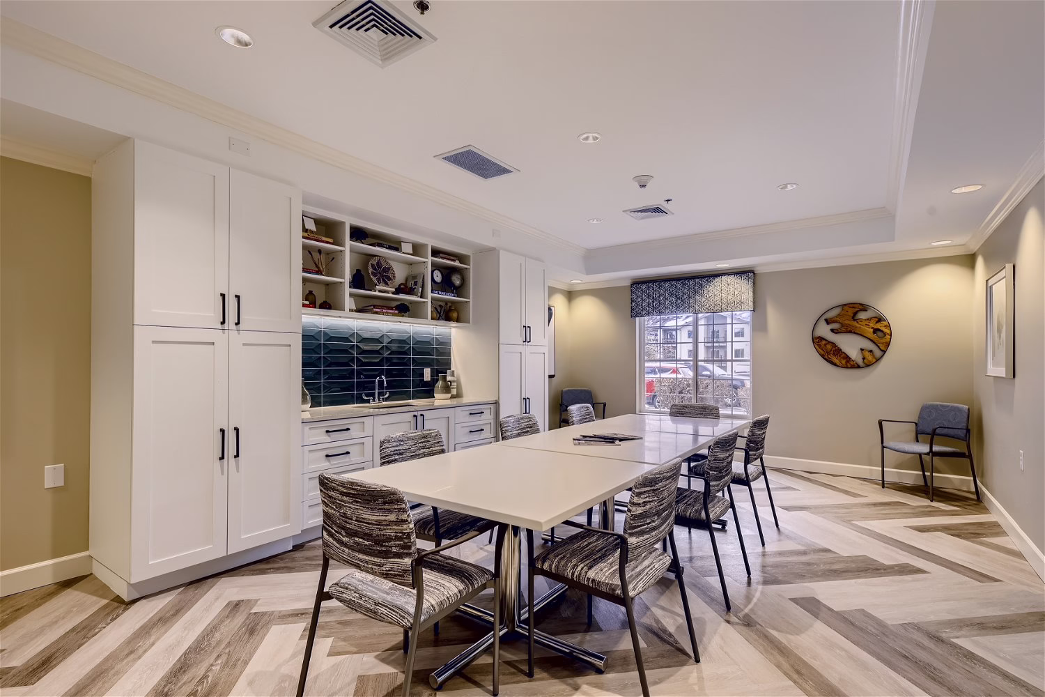 Modern meeting room with a long table and six chairs, built-in cabinets, open shelves with decor, parquet flooring, and a large window with city view. Neutral tones and contemporary art create a stylish, inviting space.