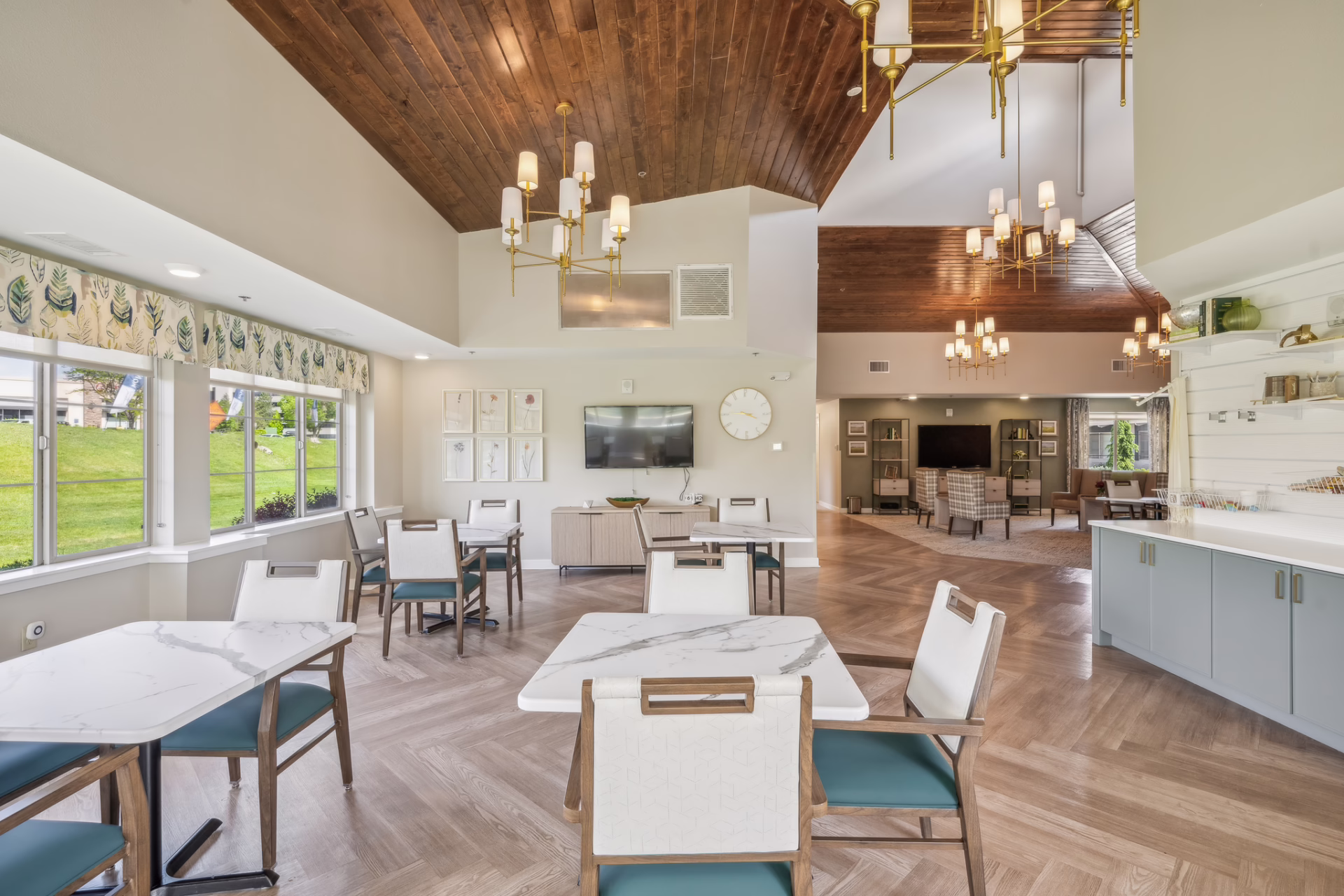 Bright senior living gathering space with a wood slat ceiling and expansive windows overlooking the lawn, by Aneka Interiors